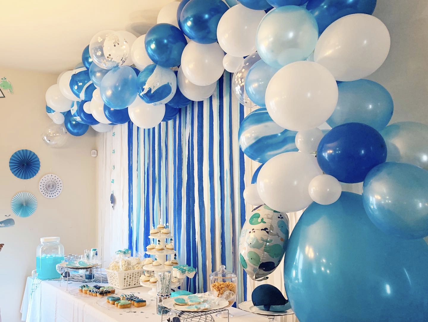 Party table decorated with blue and white balloons, striped blue curtains, and a dessert display.