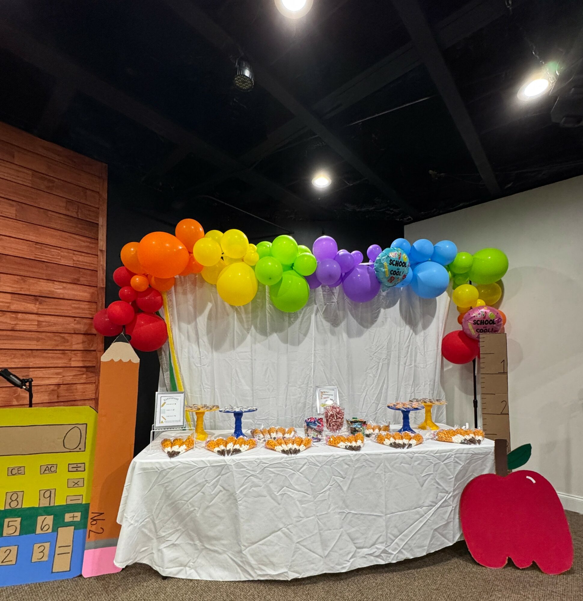 Party setup with colorful balloons, a table with a white cloth, and a large red apple decoration.