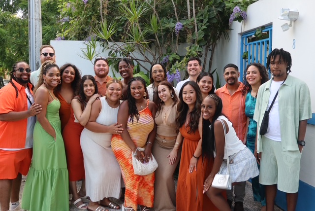 Group of people smiling outdoors, standing in front of a white wall with greenery and purple flowers.
