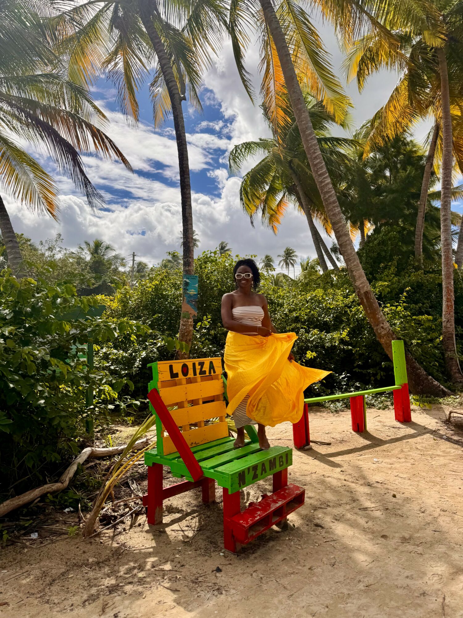 Woman in yellow dress sitting on a colorful bench outdoors with palm trees and blue sky in background.