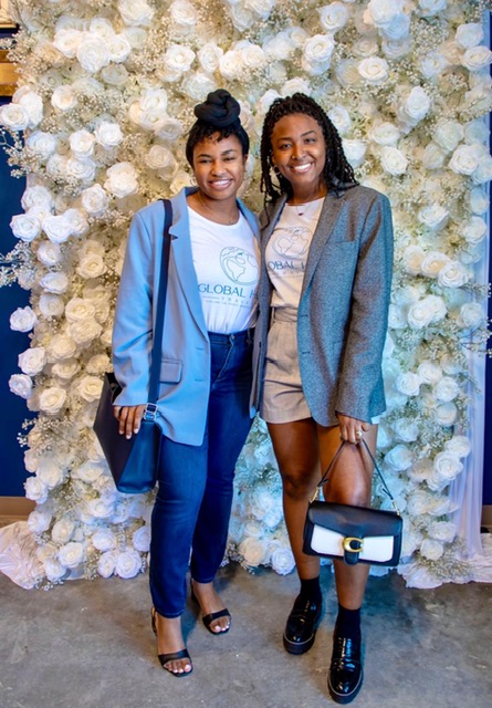 Two women stand in front of a white floral backdrop, smiling, with one holding a handbag and the other with a backpack.