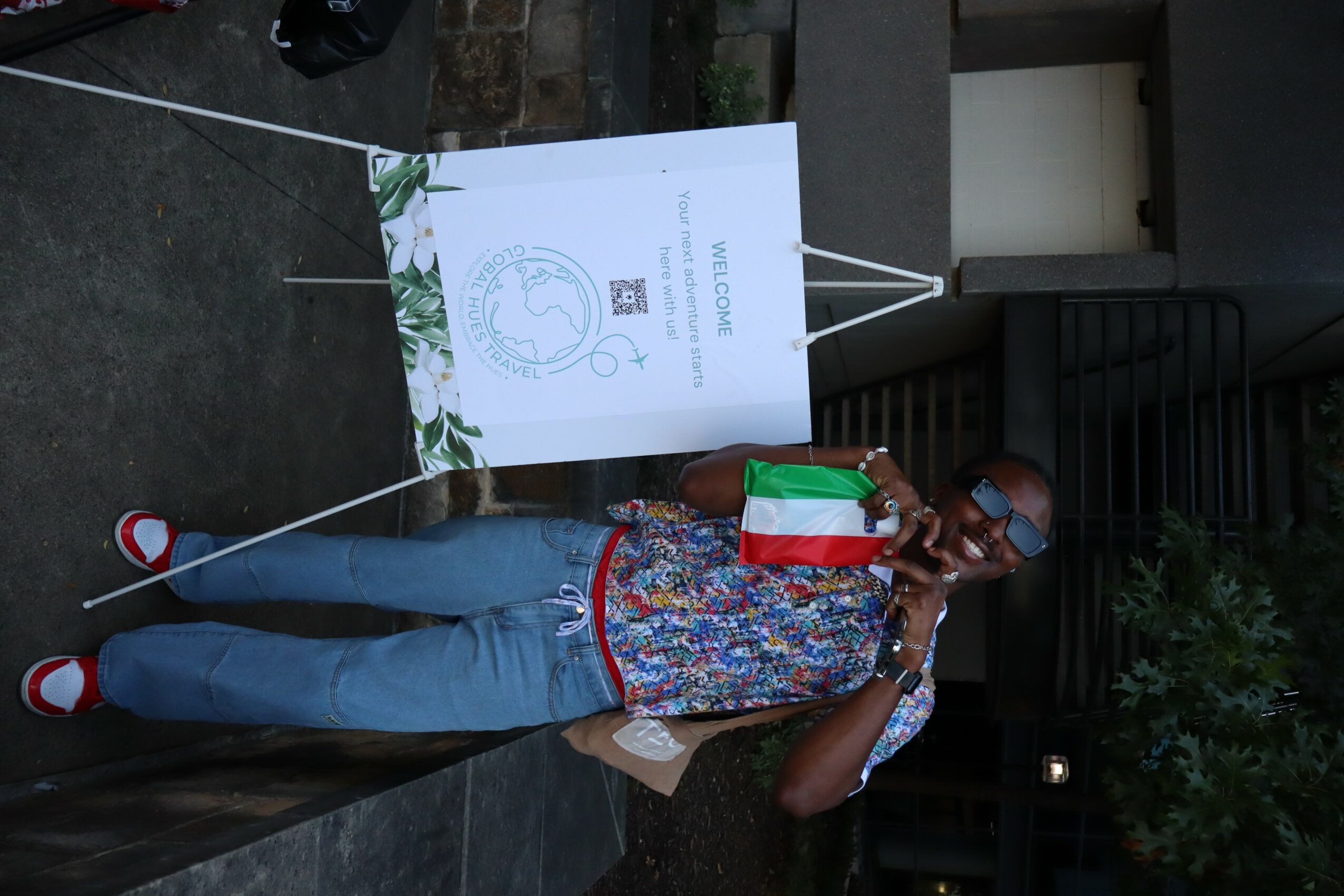 Person standing outdoors near a sign, wearing sunglasses, a colorful shirt, jeans, and red shoes, holding a drink.