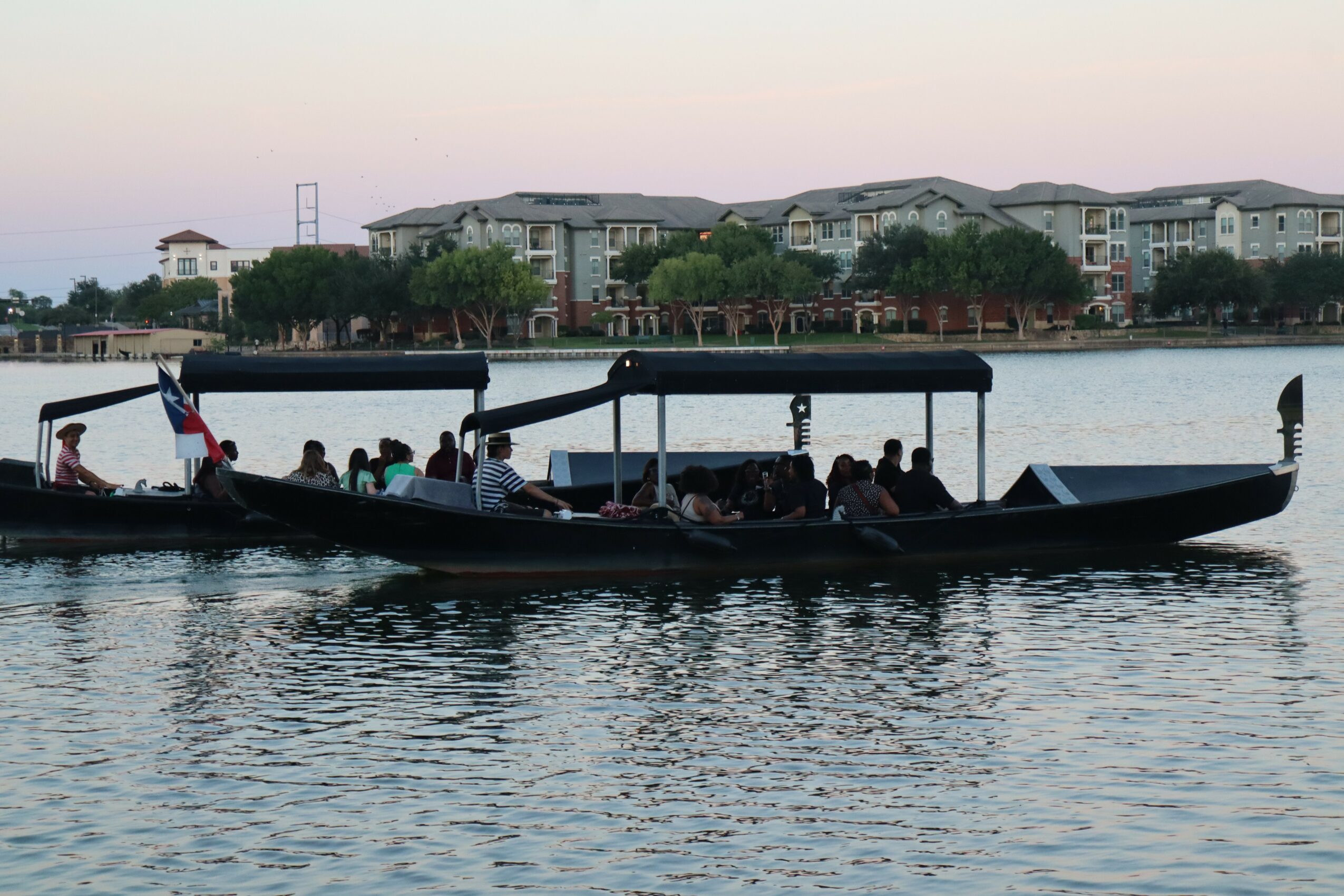 Long boat with people on board floating on water, buildings and trees in background, evening sky.
