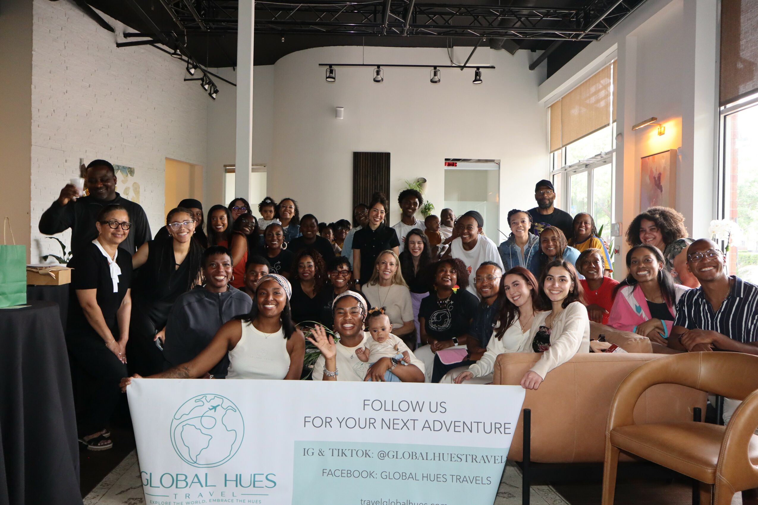 Group of diverse people gathered indoors, some seated and some standing, in a room with large windows and a white wall.
