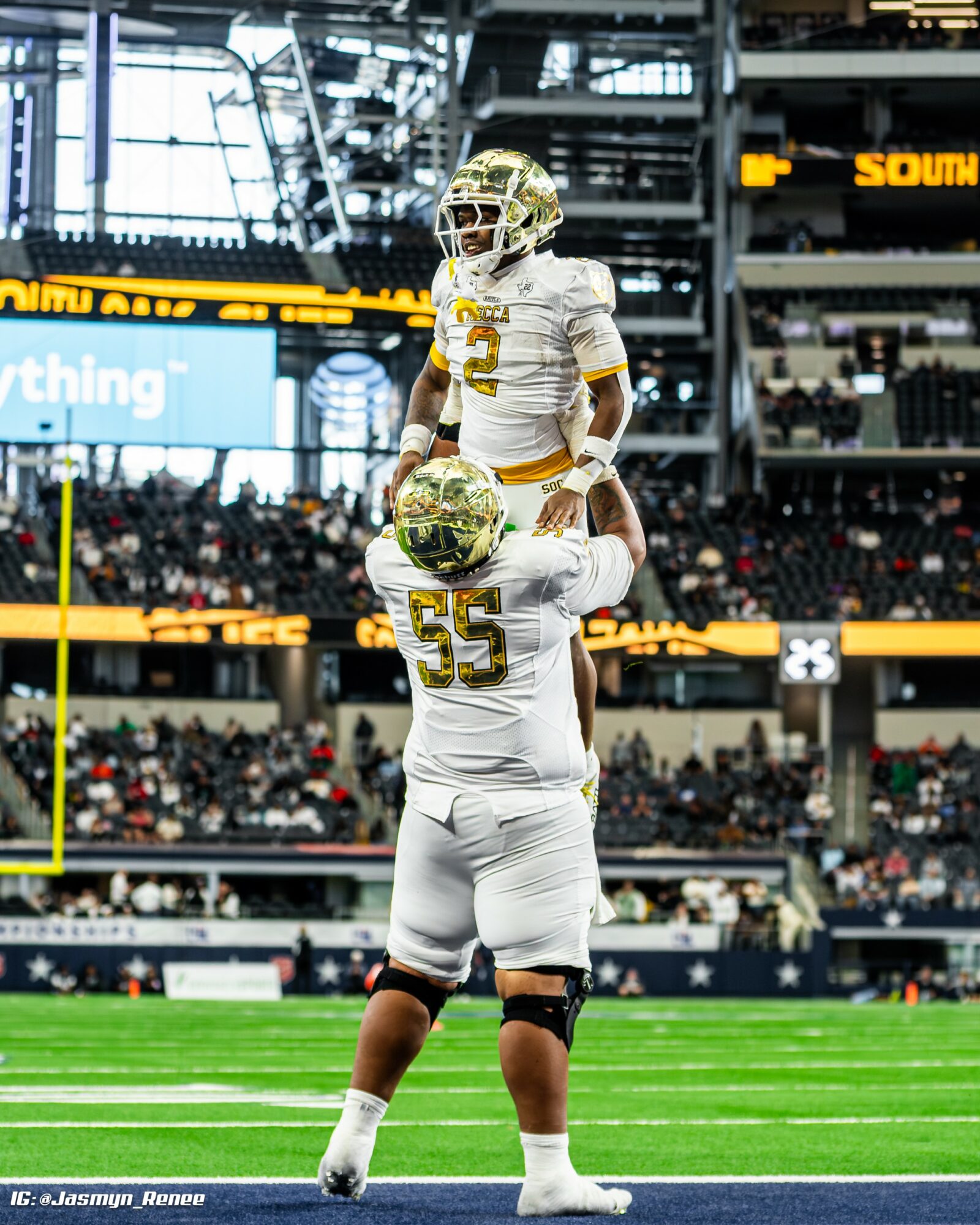 Two football players in white uniforms on field, one holding a helmet, stadium with spectators in background.
