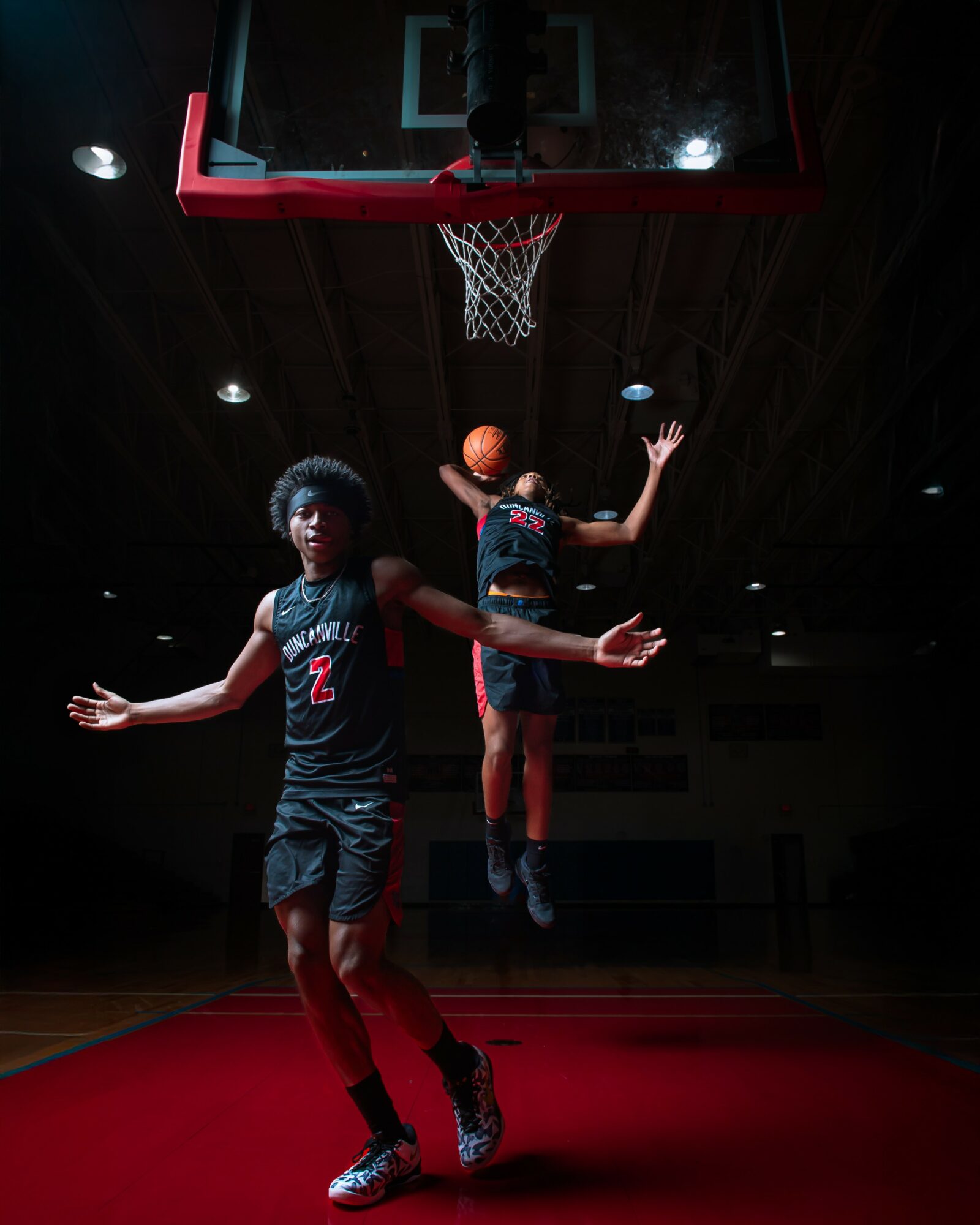 Two basketball players jump towards the hoop in a dark indoor gym, one holding a basketball, with a red court floor.