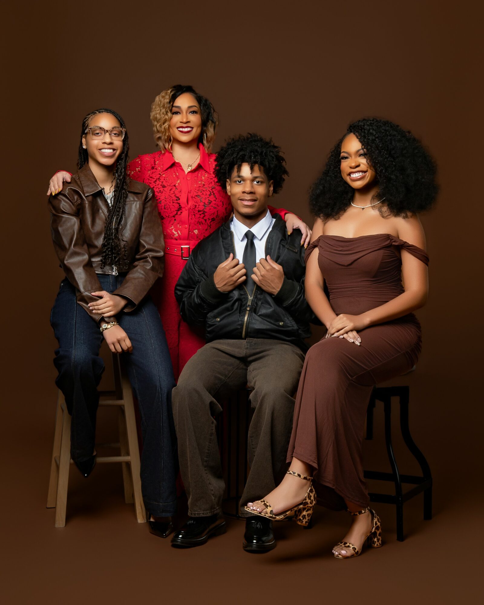 Four people, three women and one man, pose together against a plain brown background, smiling and dressed stylishly.