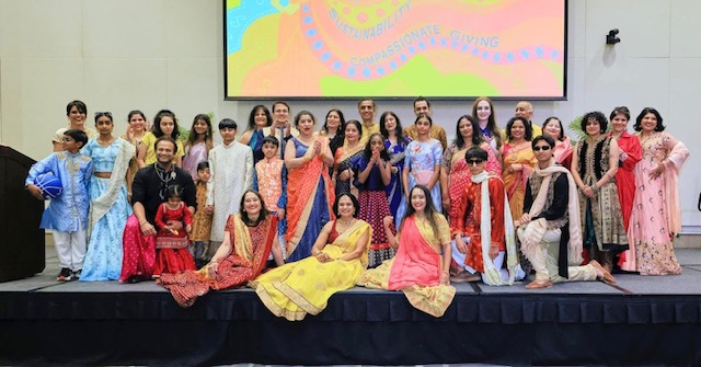 Group of women and children in colorful traditional dresses on stage, some sitting and some standing, with a screen behind.