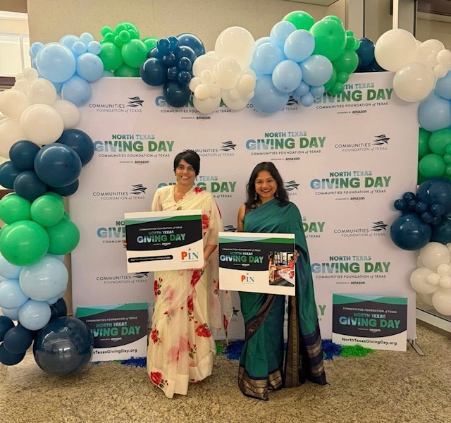 Two women in traditional attire standing in front of a decorated backdrop with balloons, holding large gift cards.