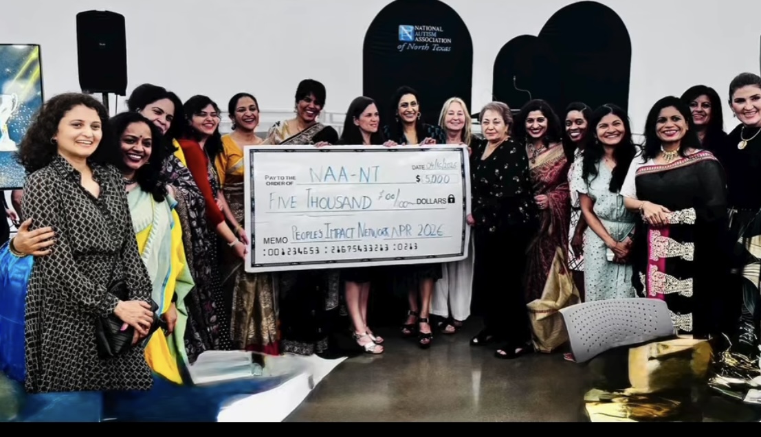 Group of women holding a large check, smiling indoors, some wearing traditional dresses, in a room with black and white decor.