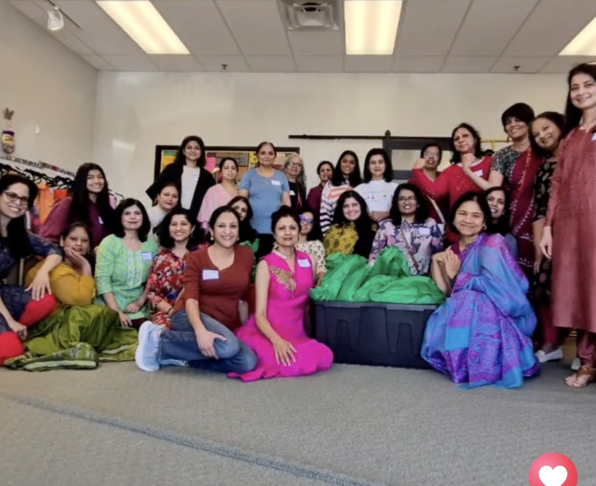 Group of women gathered in a room, some sitting and some standing, with colorful clothing and a large green bag in the center.