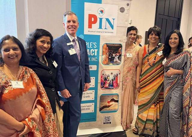 Group of seven people standing indoors, smiling, with a banner behind them displaying the PIN logo and images.