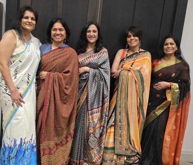 Five women standing together, smiling, wearing colorful sarees, in front of a dark wall.