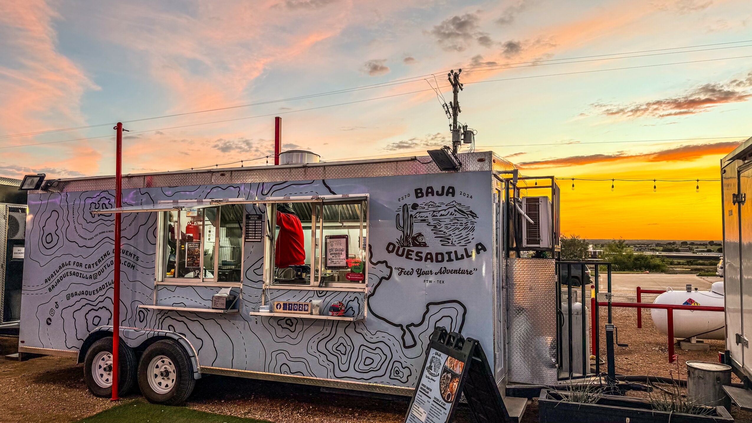 Food truck with open window, person inside, outdoor setting during sunset, sky with clouds, trees in background.