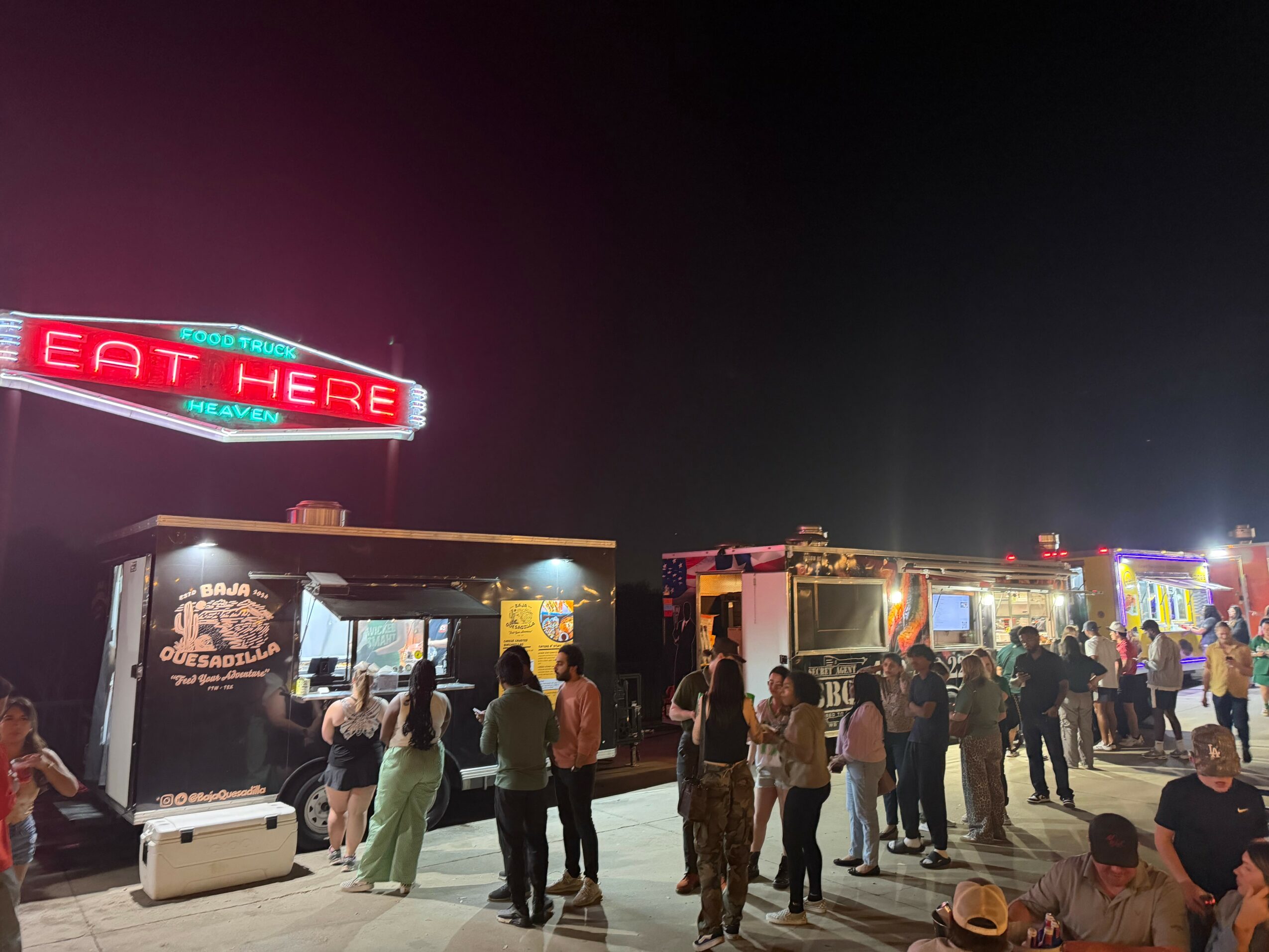Night scene with food trucks and people gathered outside, illuminated by neon and string lights.