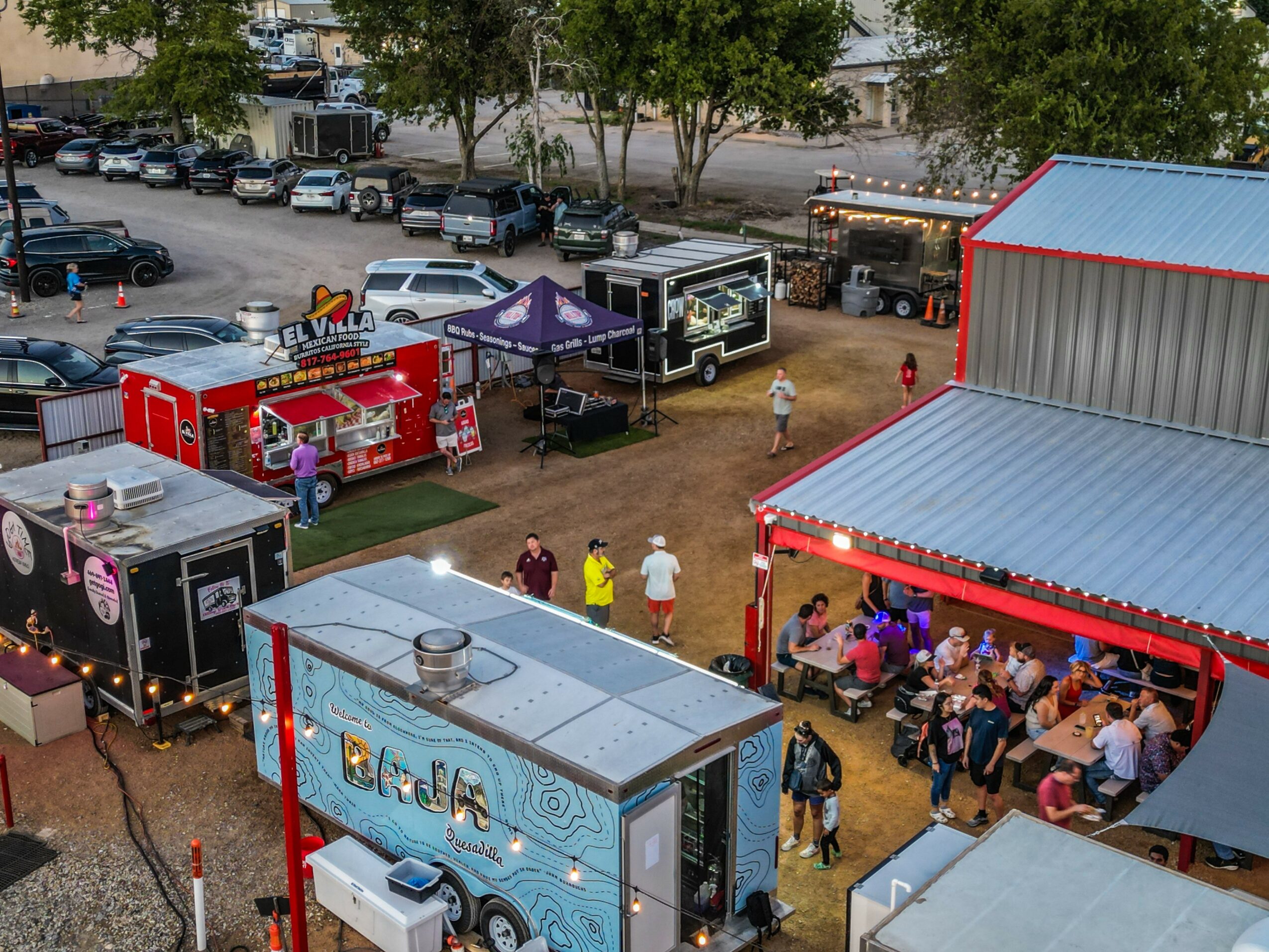Outdoor event with food trucks, tents, and people gathered under a red-roofed shelter.
