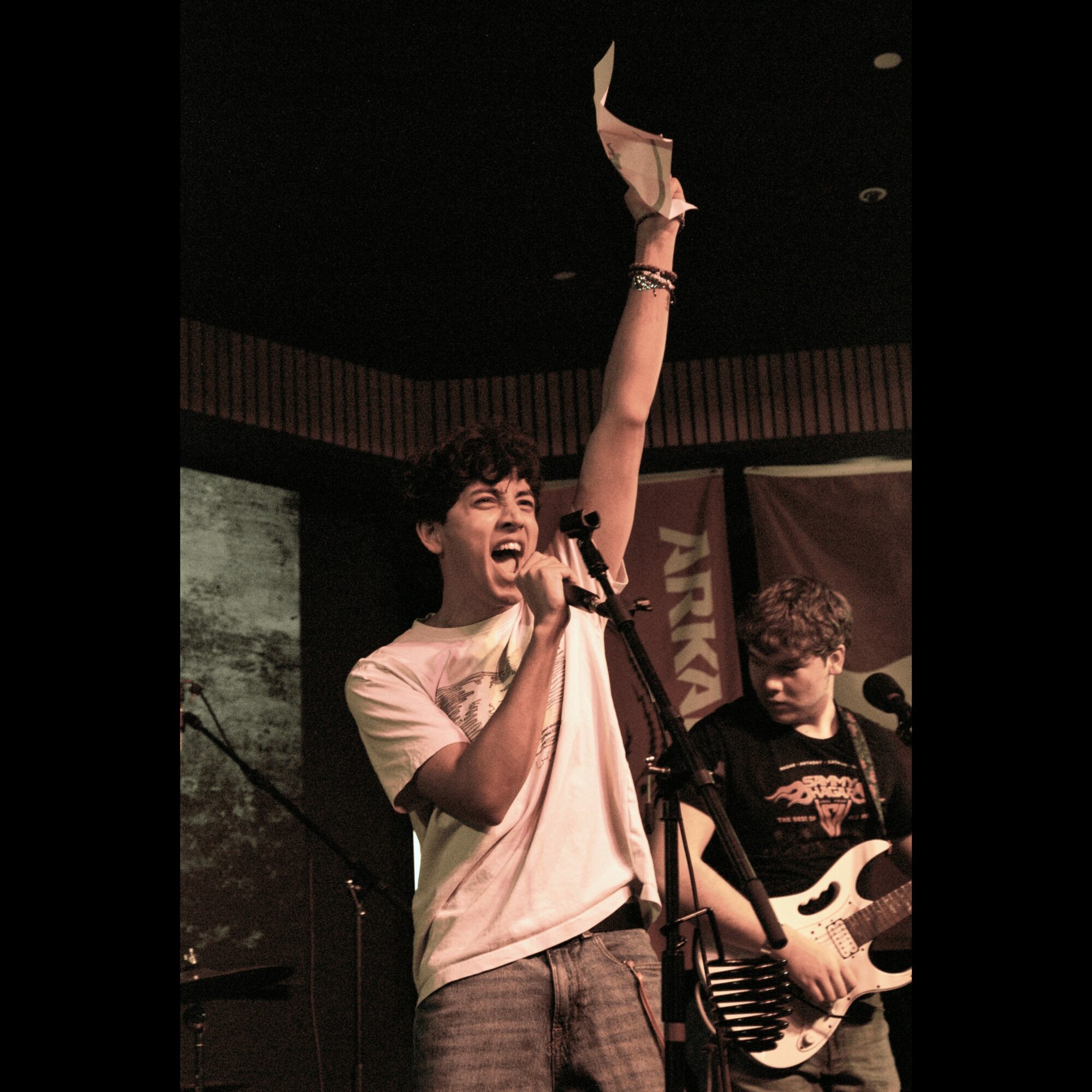 Person singing into microphone with arm raised, another person playing guitar in background, stage setting.
