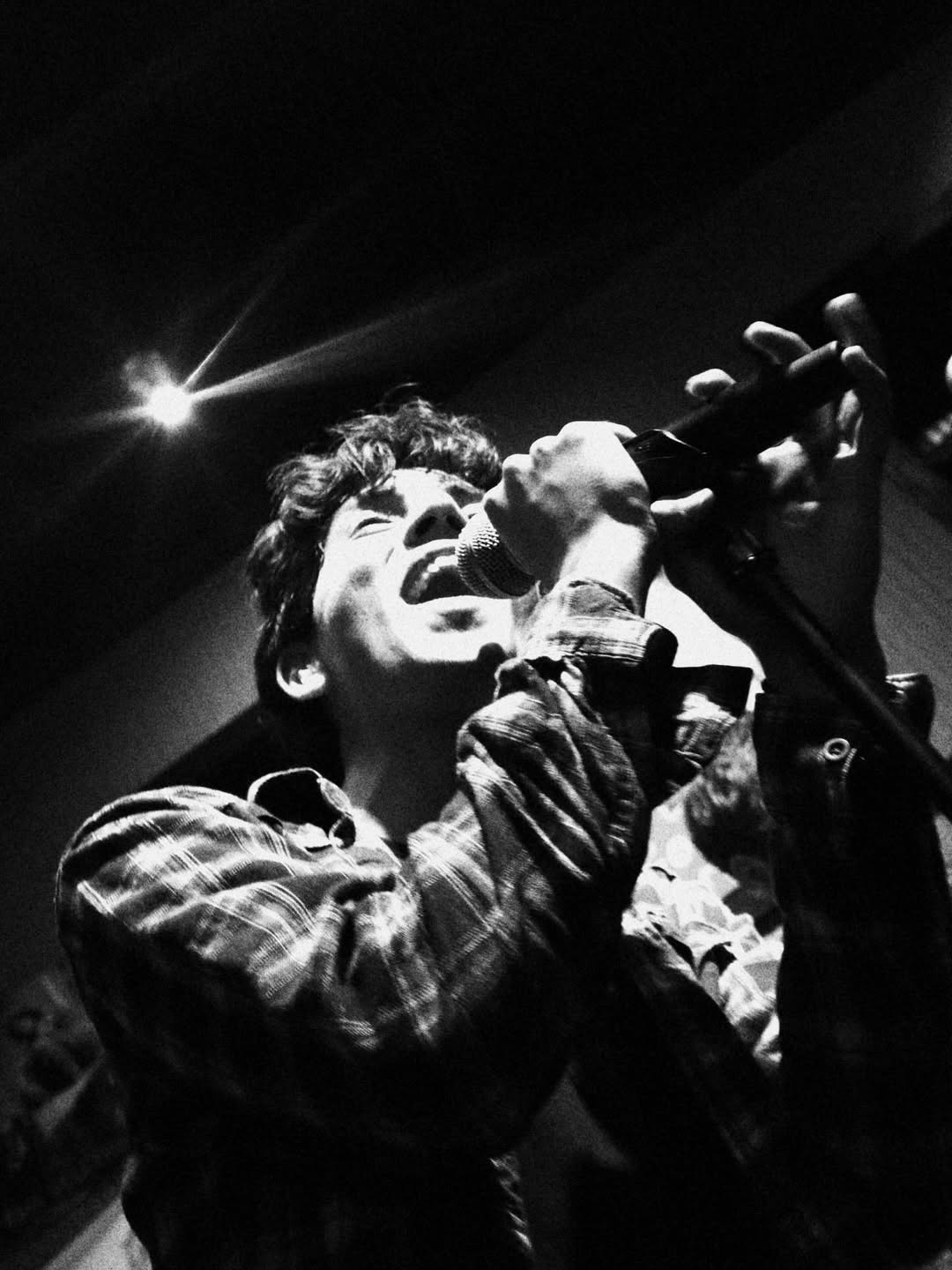 Person singing into a microphone on stage with dramatic lighting, wearing a checkered shirt.