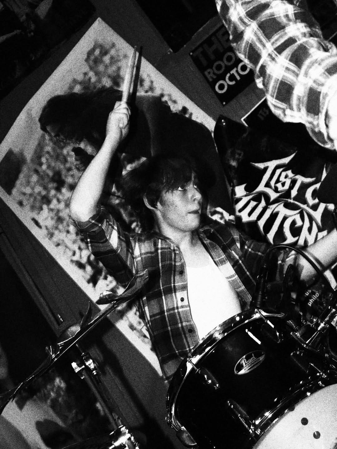 Woman playing drums with drumsticks, wearing plaid shirt, in a music venue with posters on the wall.