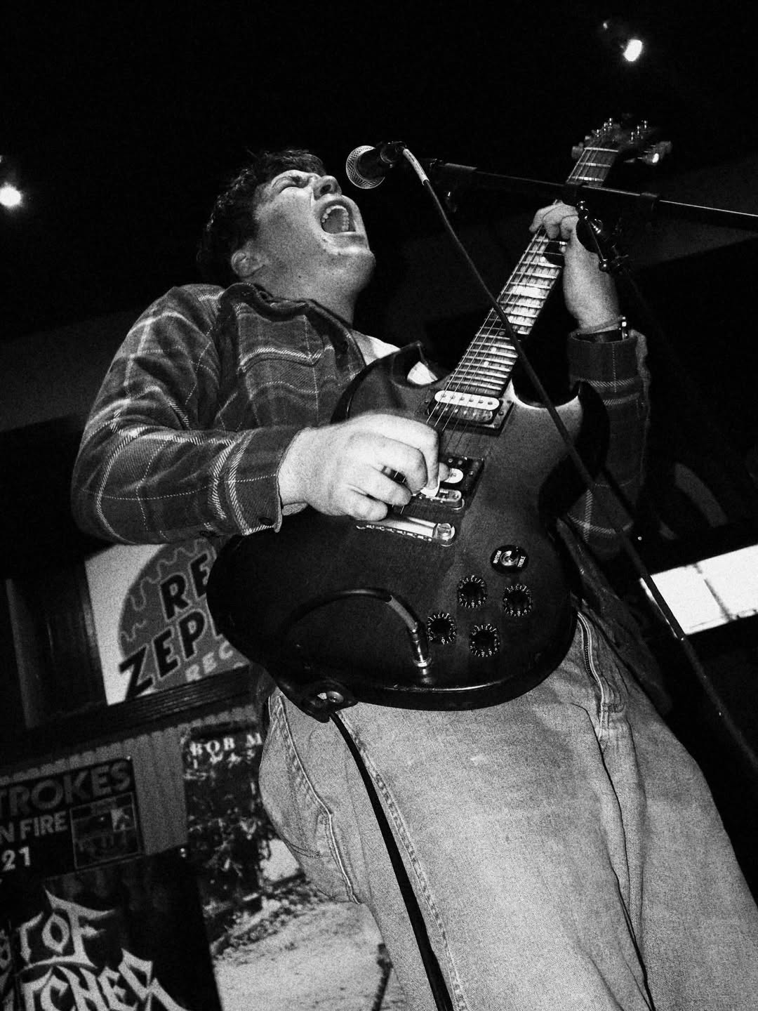 Person singing passionately while playing an electric guitar on stage, with a microphone in front, in black and white.