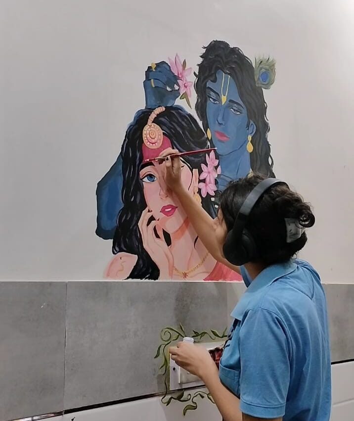Person wearing headphones and a blue shirt painting a mural of two women with flowers and jewelry on a wall.
