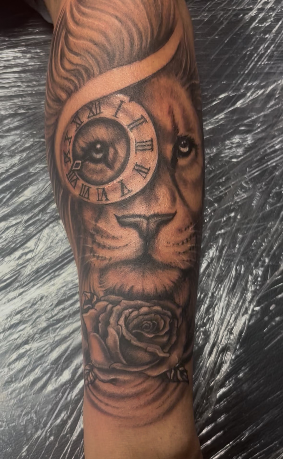 Tattoo of a lion's face with a clock and a rose on the arm.