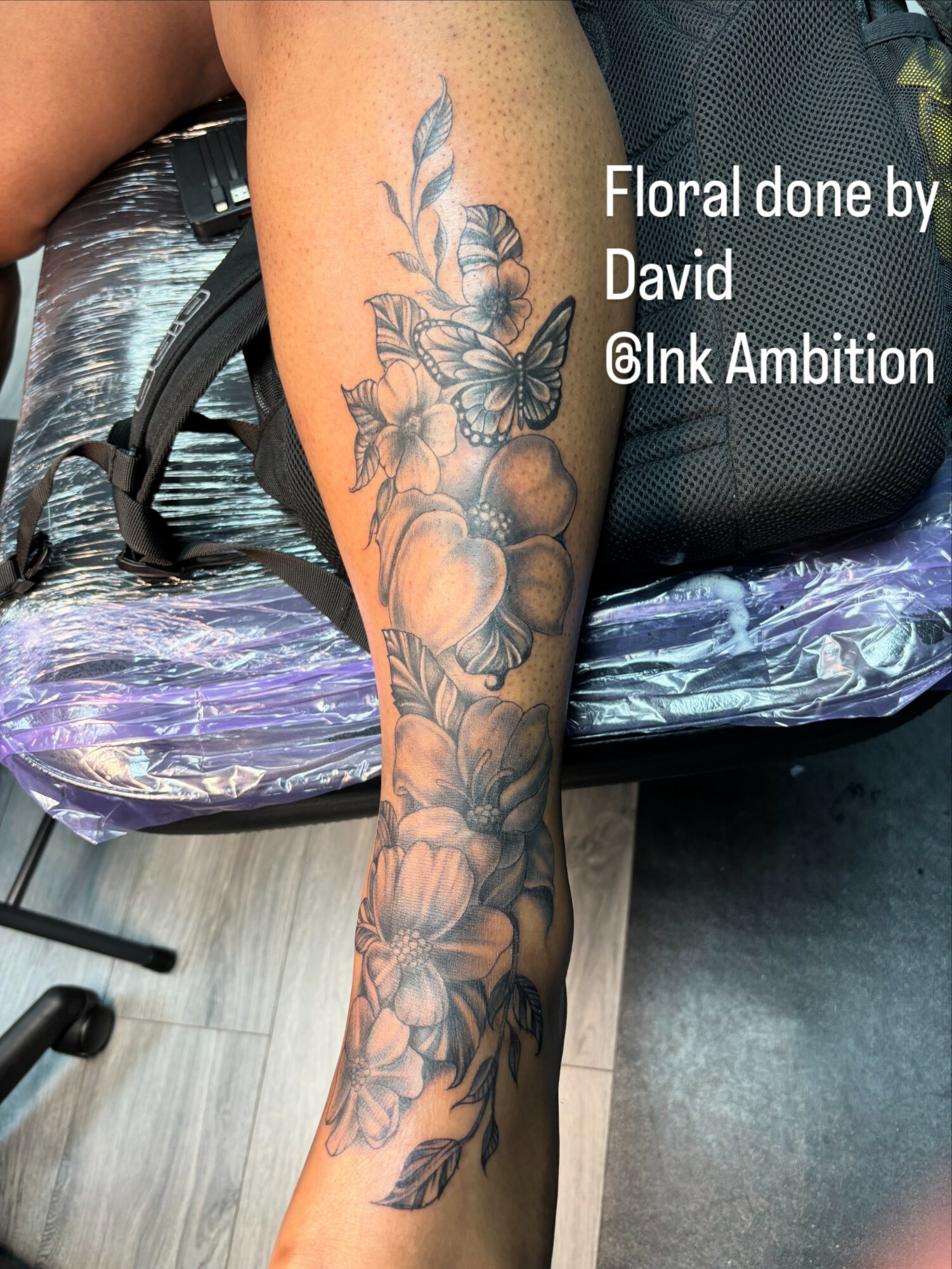 Tattoo of flowers and leaves on person's leg, with text overlay about floral tattoo by David @Ink Ambition.