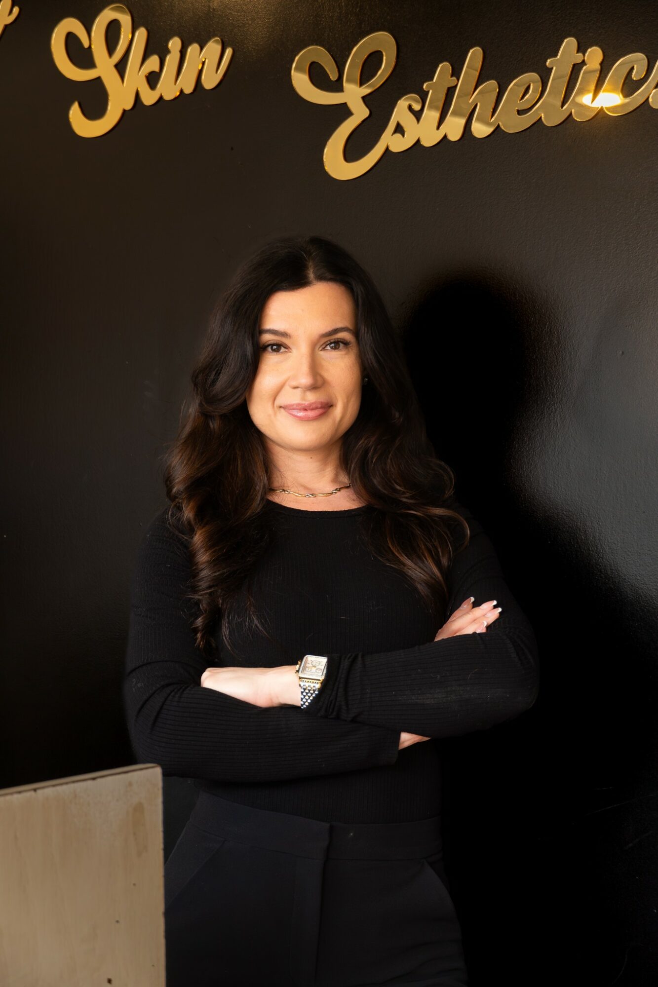 Woman with long dark hair smiling, standing with arms crossed in front of a black wall with gold lettering.