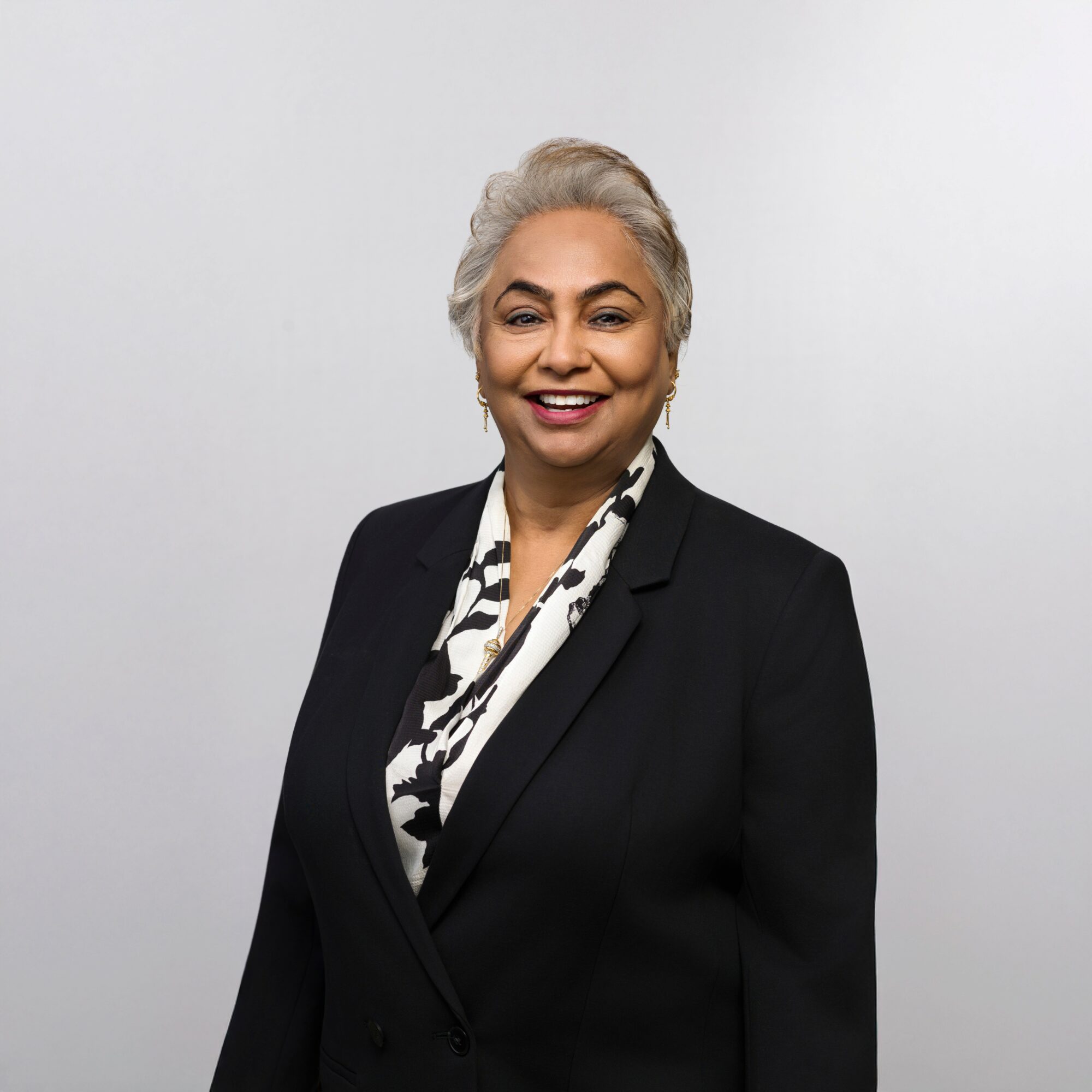 Smiling woman with gray hair wearing a black blazer and patterned blouse against a plain background.