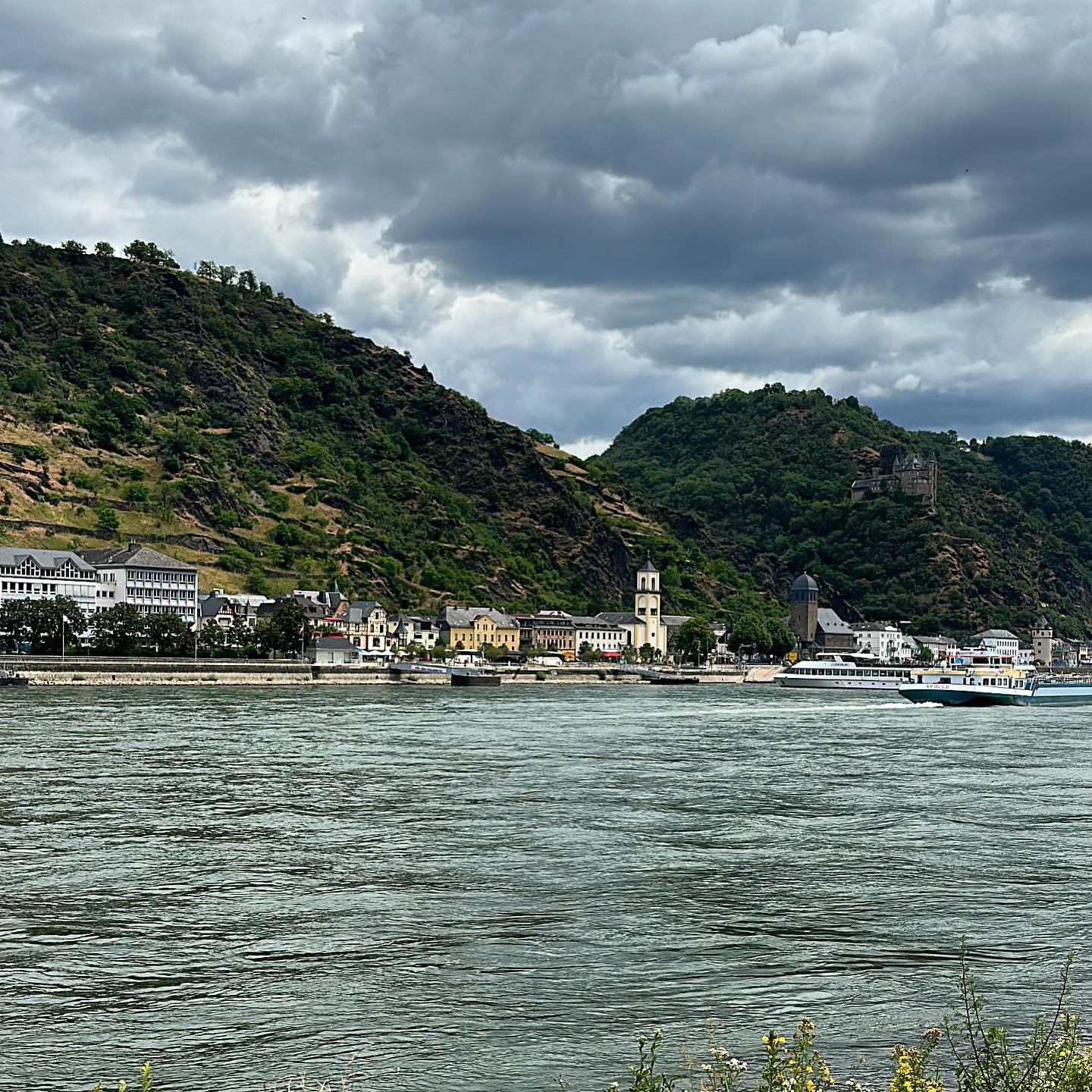 View of a river with a town and green hills in the background under cloudy sky.