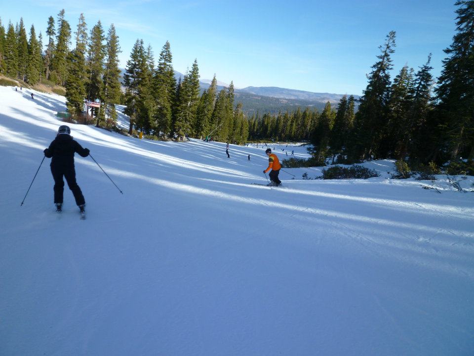 People skiing on a snow-covered slope surrounded by trees and mountains under a clear blue sky.