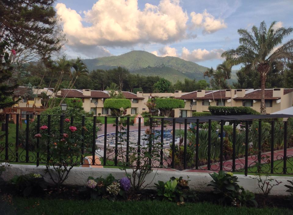 View of a residential area with houses, trees, and mountains in the background under a partly cloudy sky.