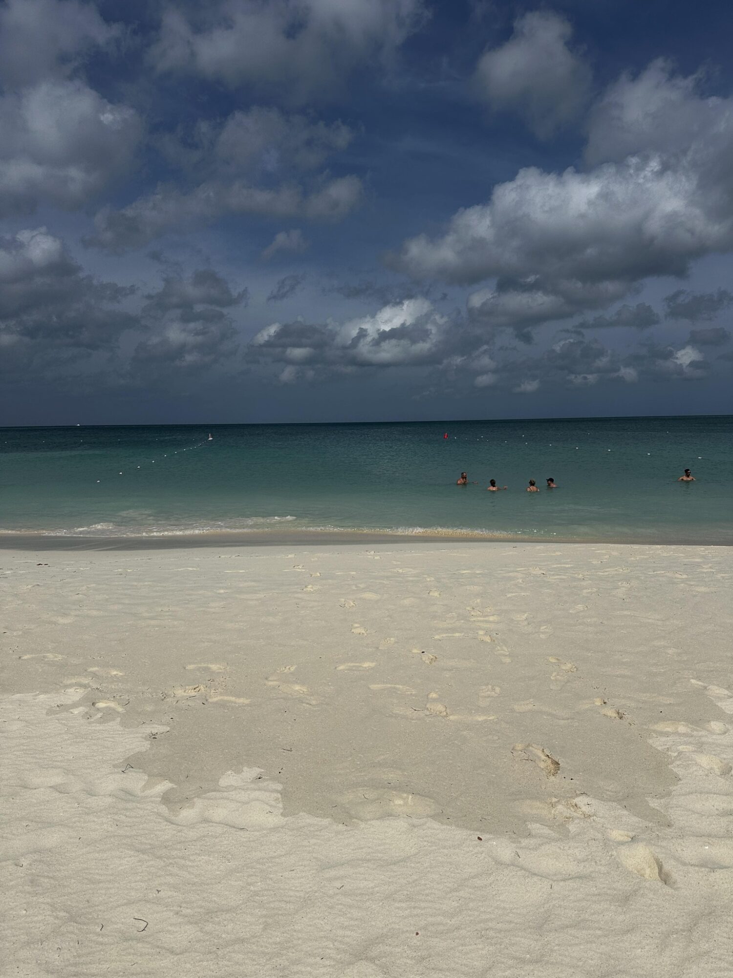 Beach with white sand, turquoise water, and a cloudy sky, with people swimming in the ocean.