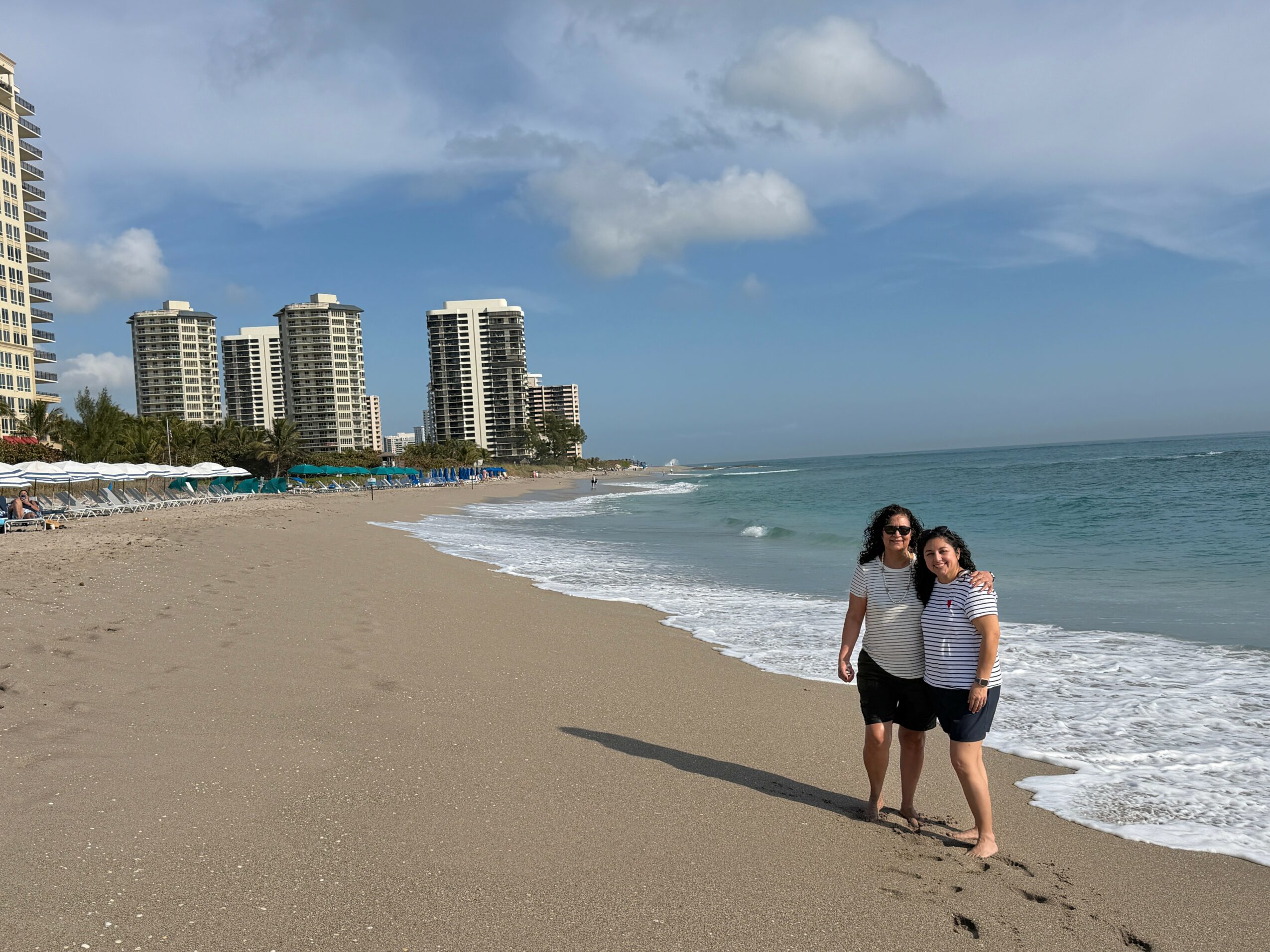 Two women stand on a sandy beach near the water, with buildings and a cloudy sky in the background.