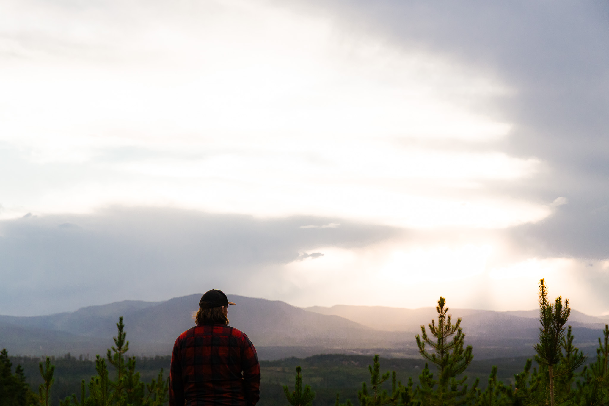 Person wearing a red and black checkered jacket and black hat standing outdoors, looking at mountains and cloudy sky.