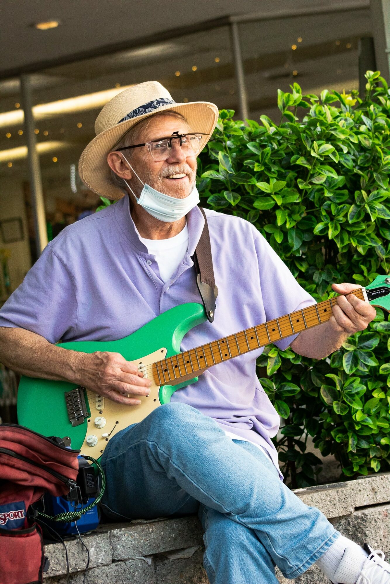 Elderly man with glasses, straw hat, and face mask playing green electric guitar outdoors near green bushes.