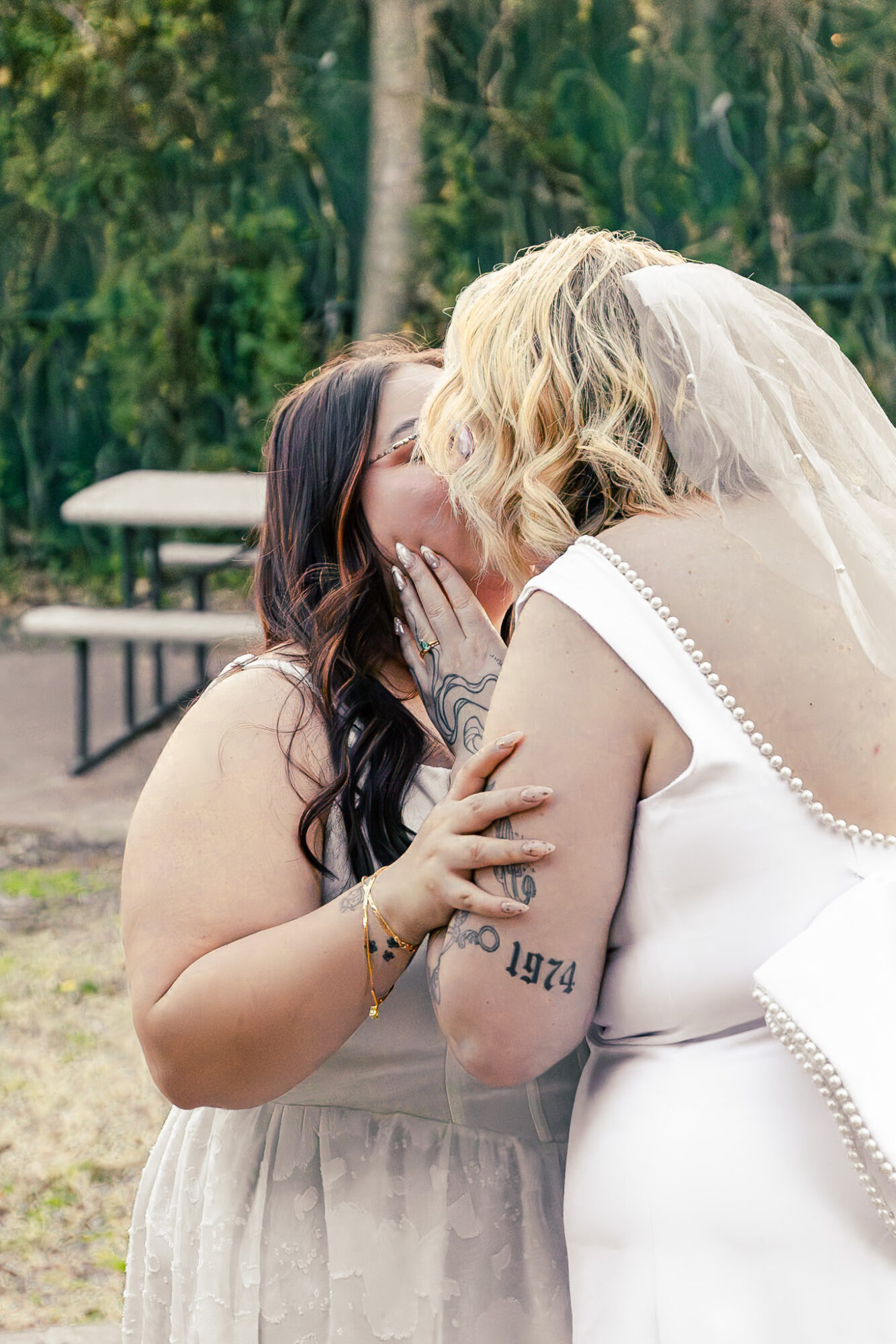 Two women share a kiss outdoors, one with dark hair and the other with blonde hair, in a natural setting.