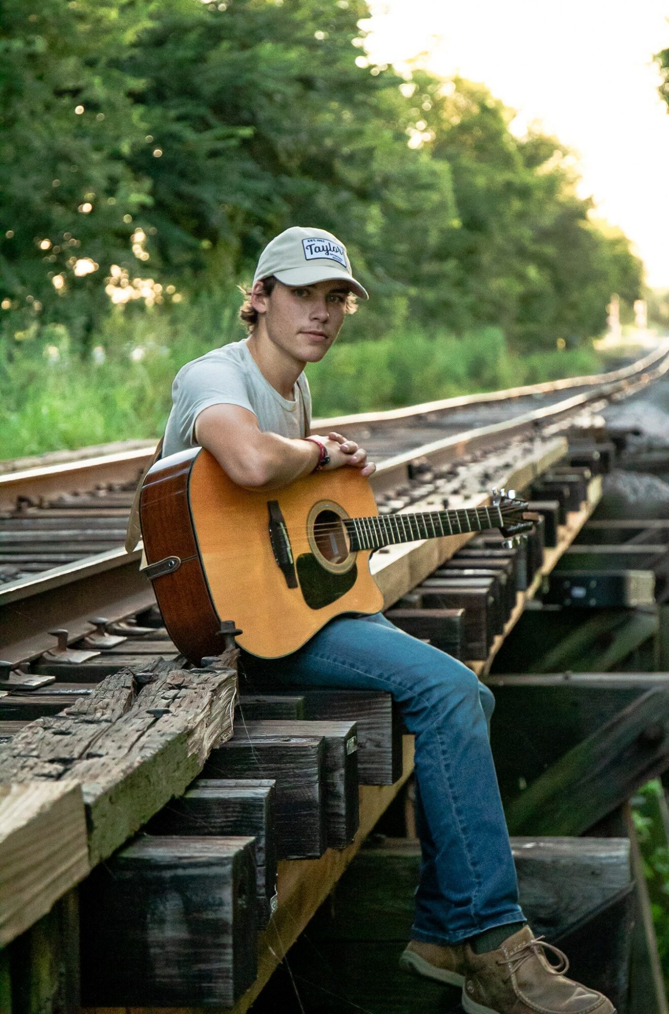 Young person sitting on a wooden railway bridge holding an acoustic guitar, surrounded by green trees and sunlight.