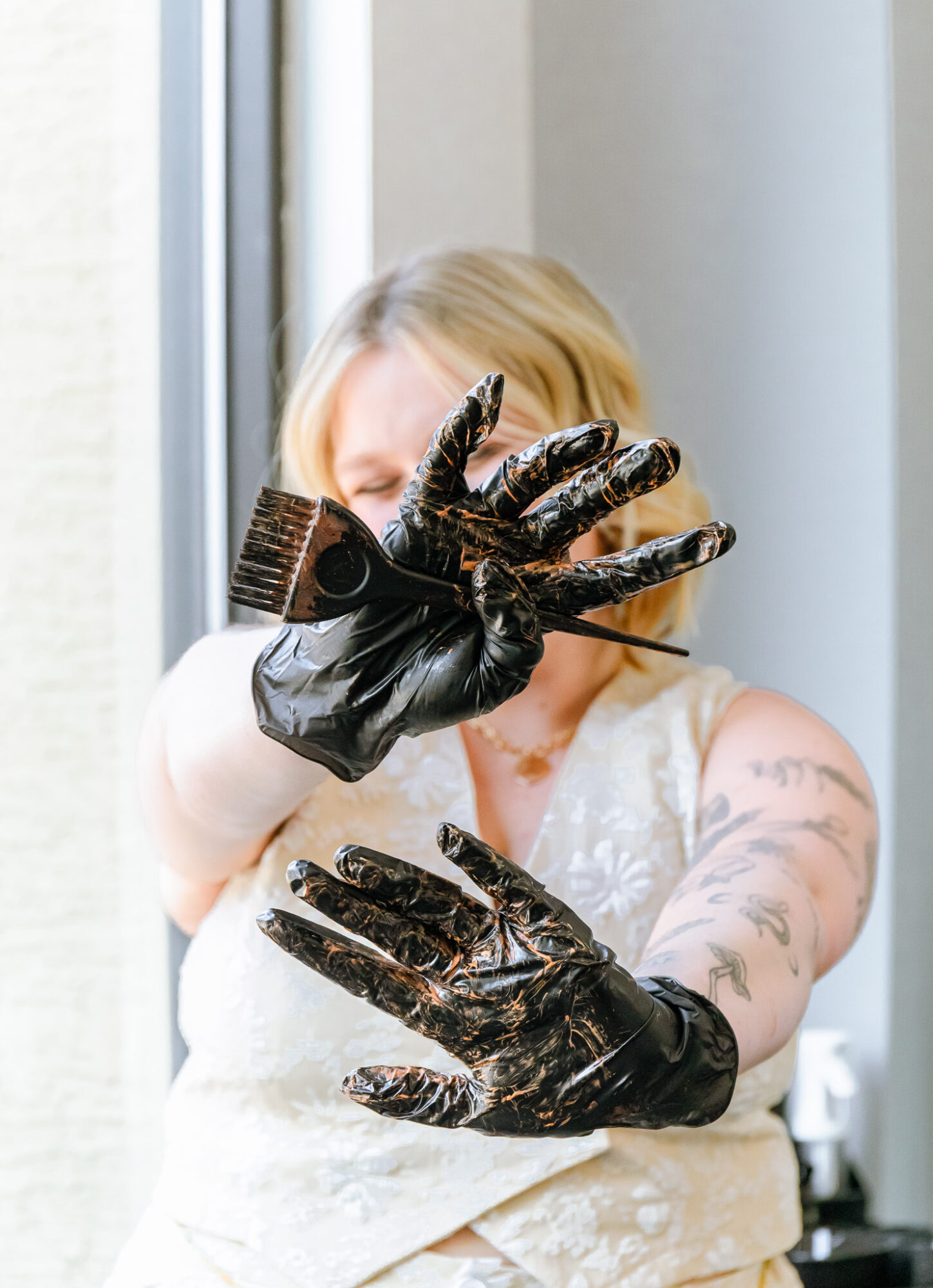 Person with blonde hair wearing black gloves, holding a dark object, standing near a window with light-colored walls.