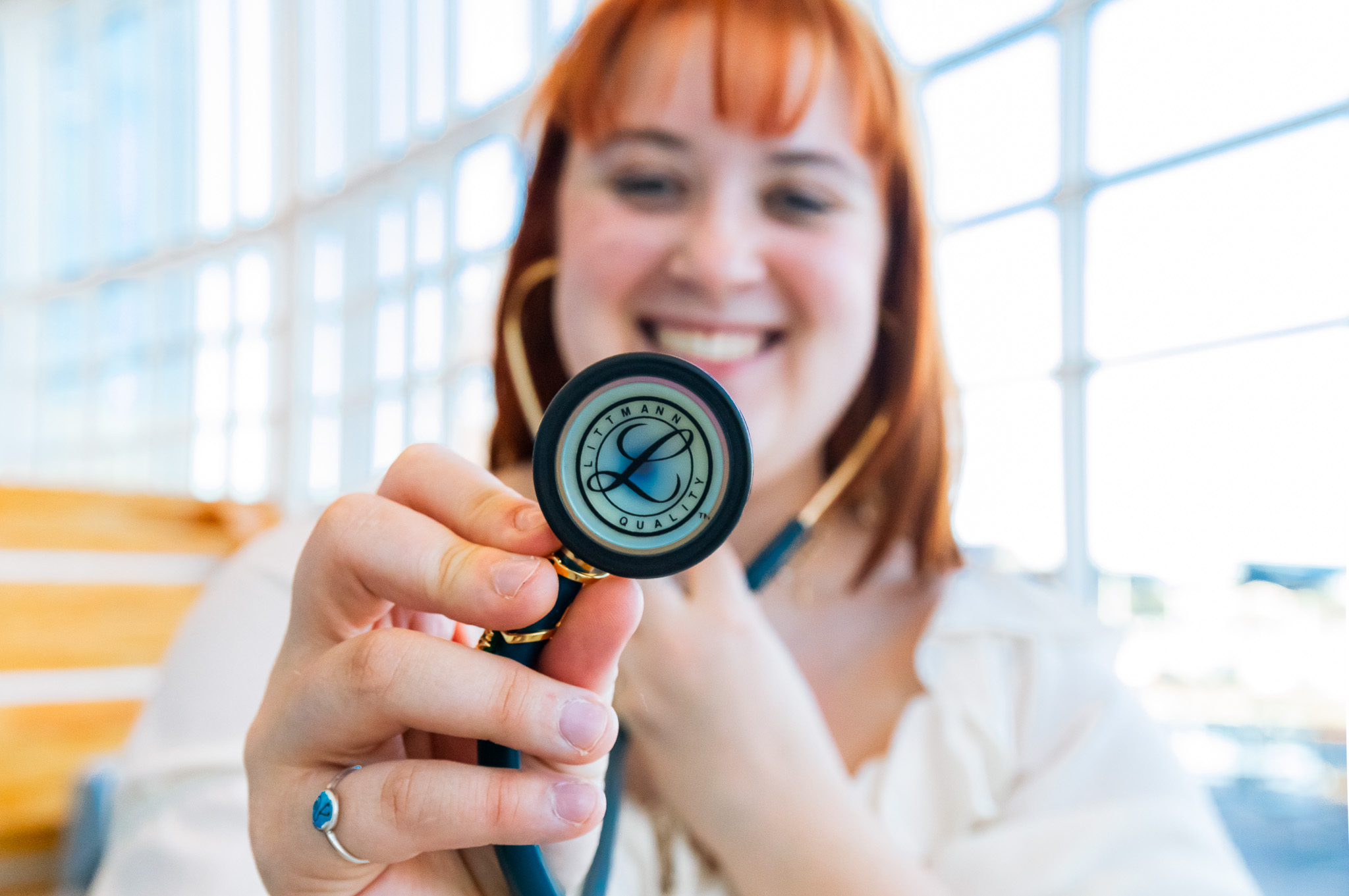 Young woman with red hair smiling, holding a stethoscope towards the camera indoors with large windows.