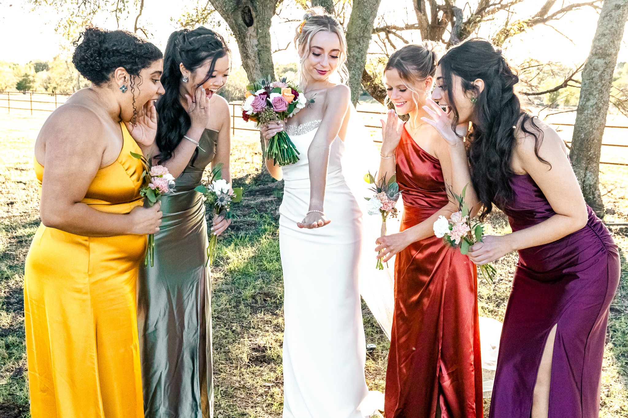 Five women in colorful dresses outdoors, one holding a bouquet, others looking at it, trees in background.