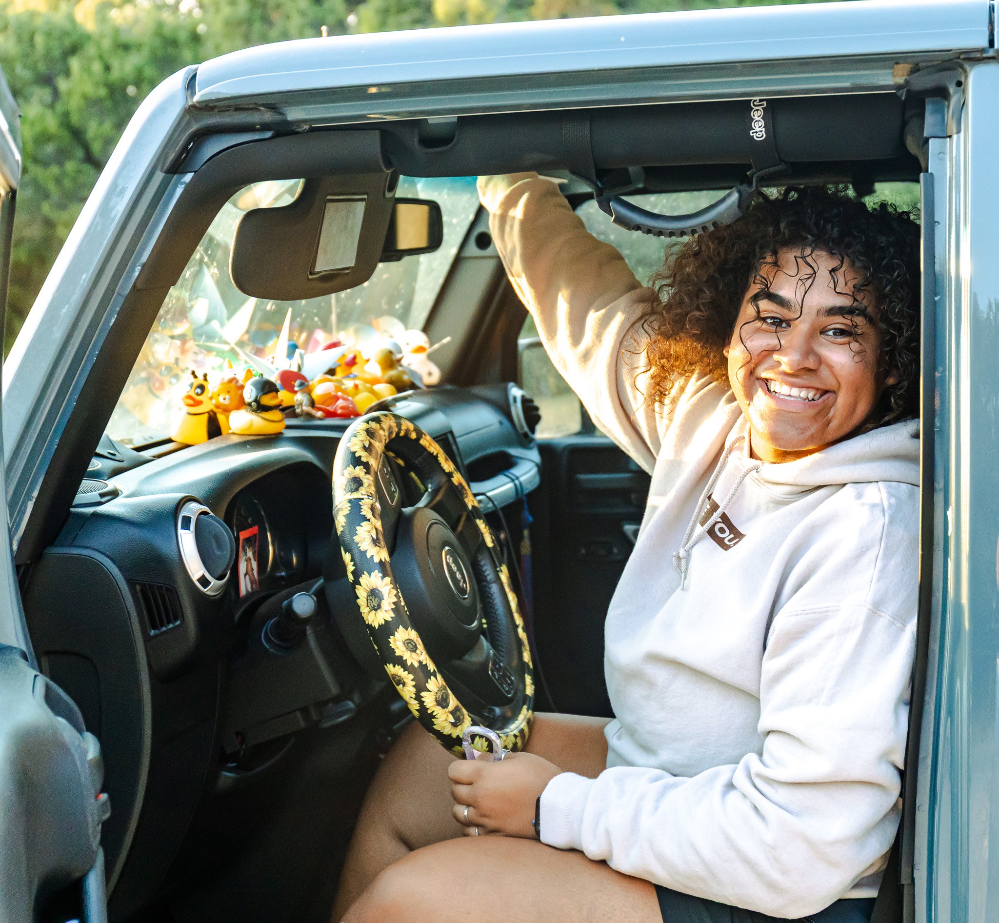 Smiling woman sitting in car with a decorated steering wheel, holding the steering wheel, with trees outside.