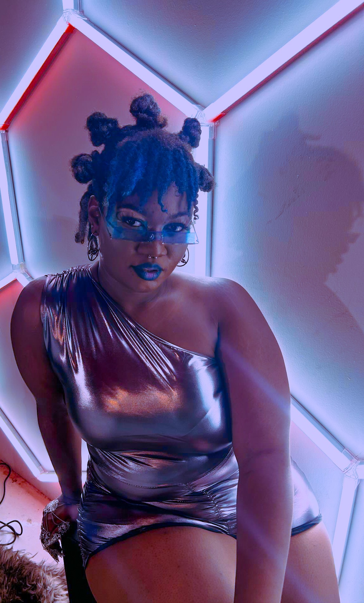 Person with styled hair, glasses, and metallic outfit in a neon-lit geometric room.