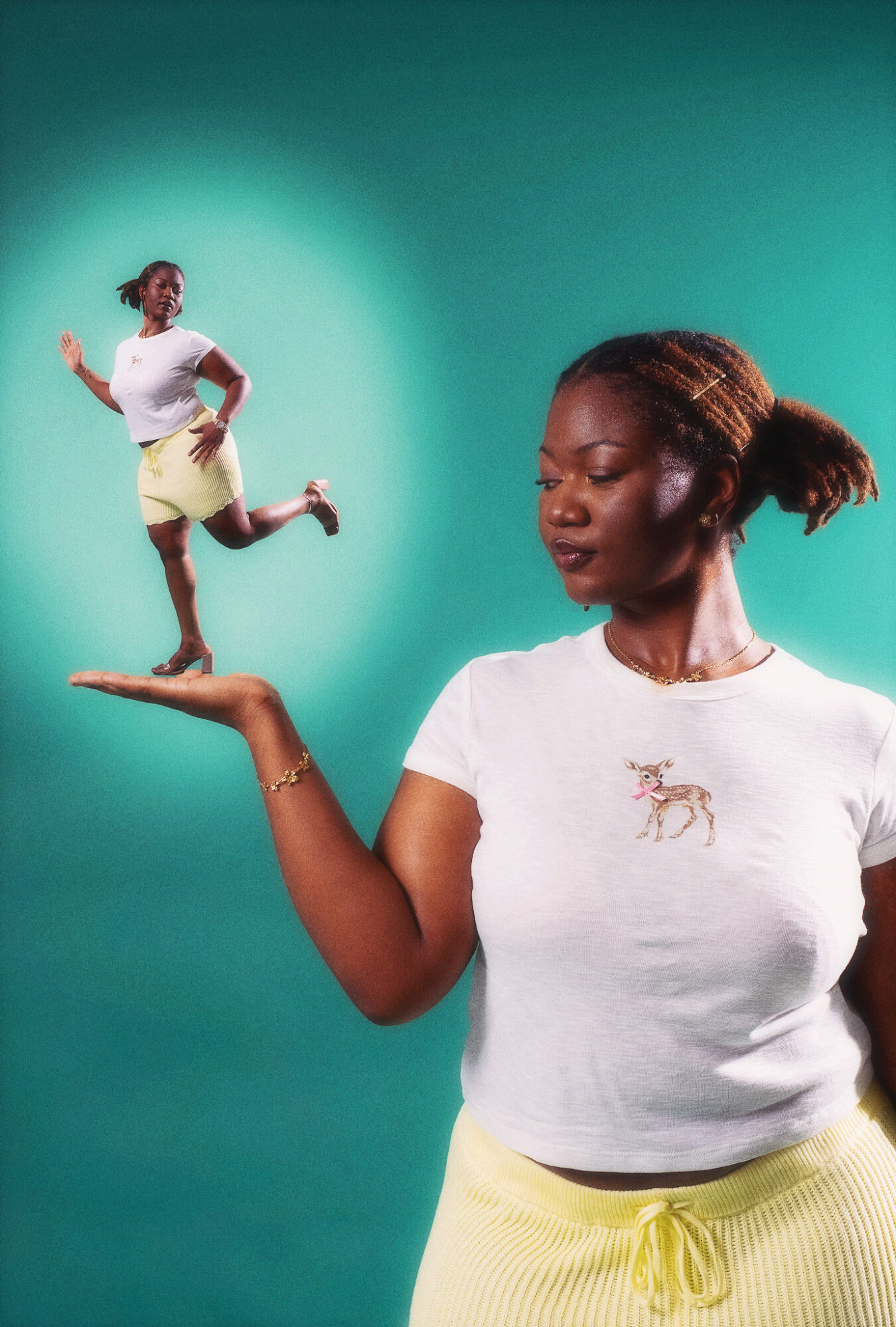 Woman with braided hair holding a small person on her hand, both wearing white shirts and yellow bottoms, against teal background.