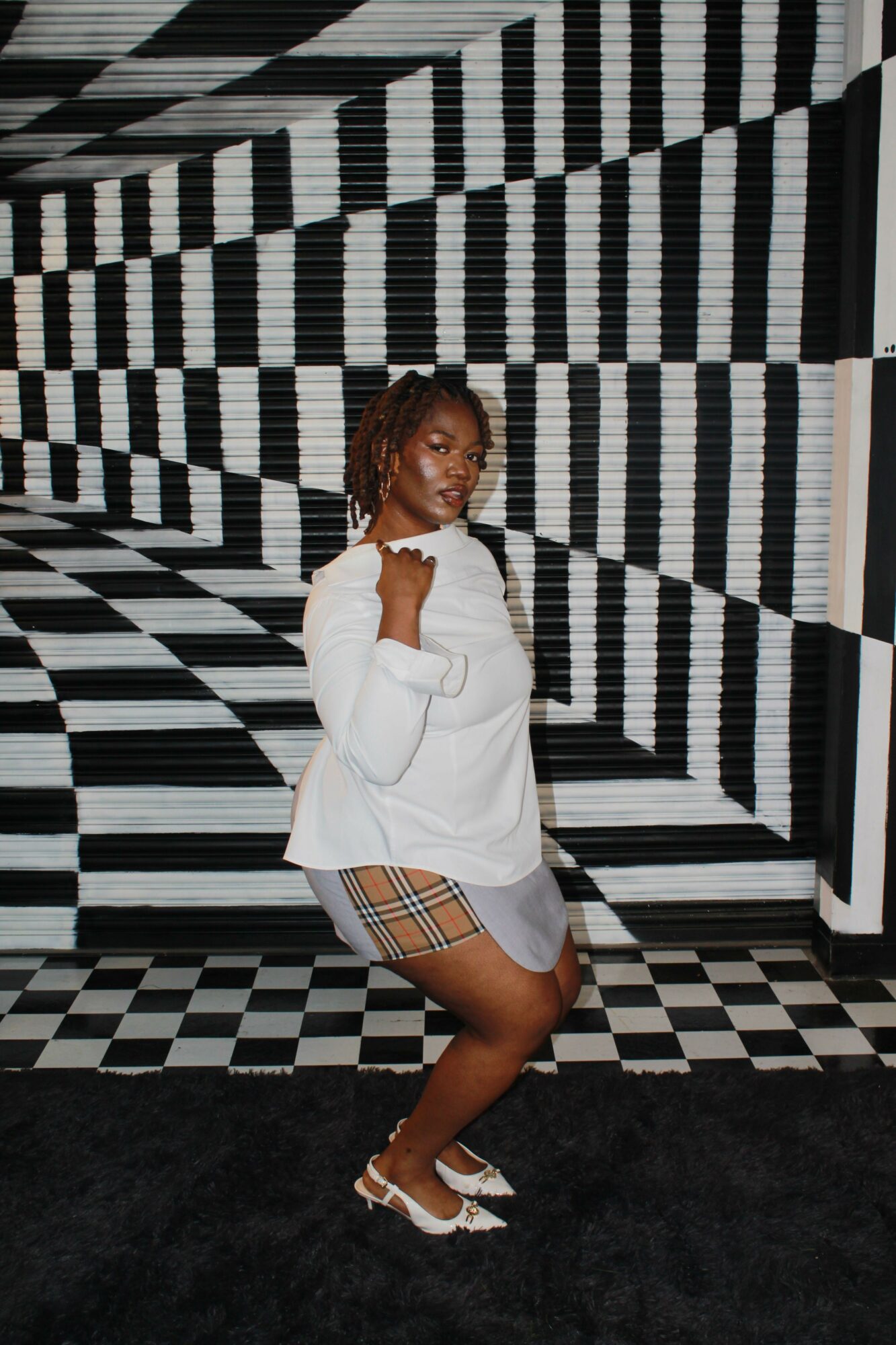 Woman with short hair in white dress and plaid shorts standing in black-and-white checkered room.