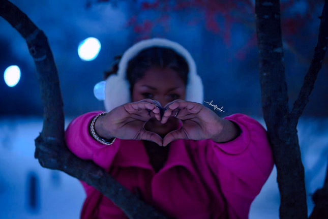Child wearing pink coat and white earmuffs making a heart shape with hands outdoors at night.