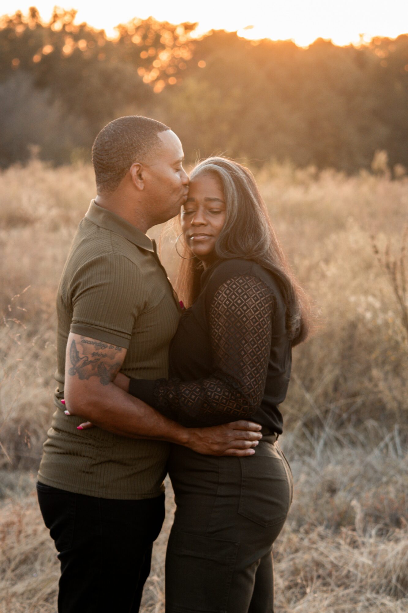 Man and woman embrace outdoors during sunset, with trees in background, eyes closed, peaceful expression.