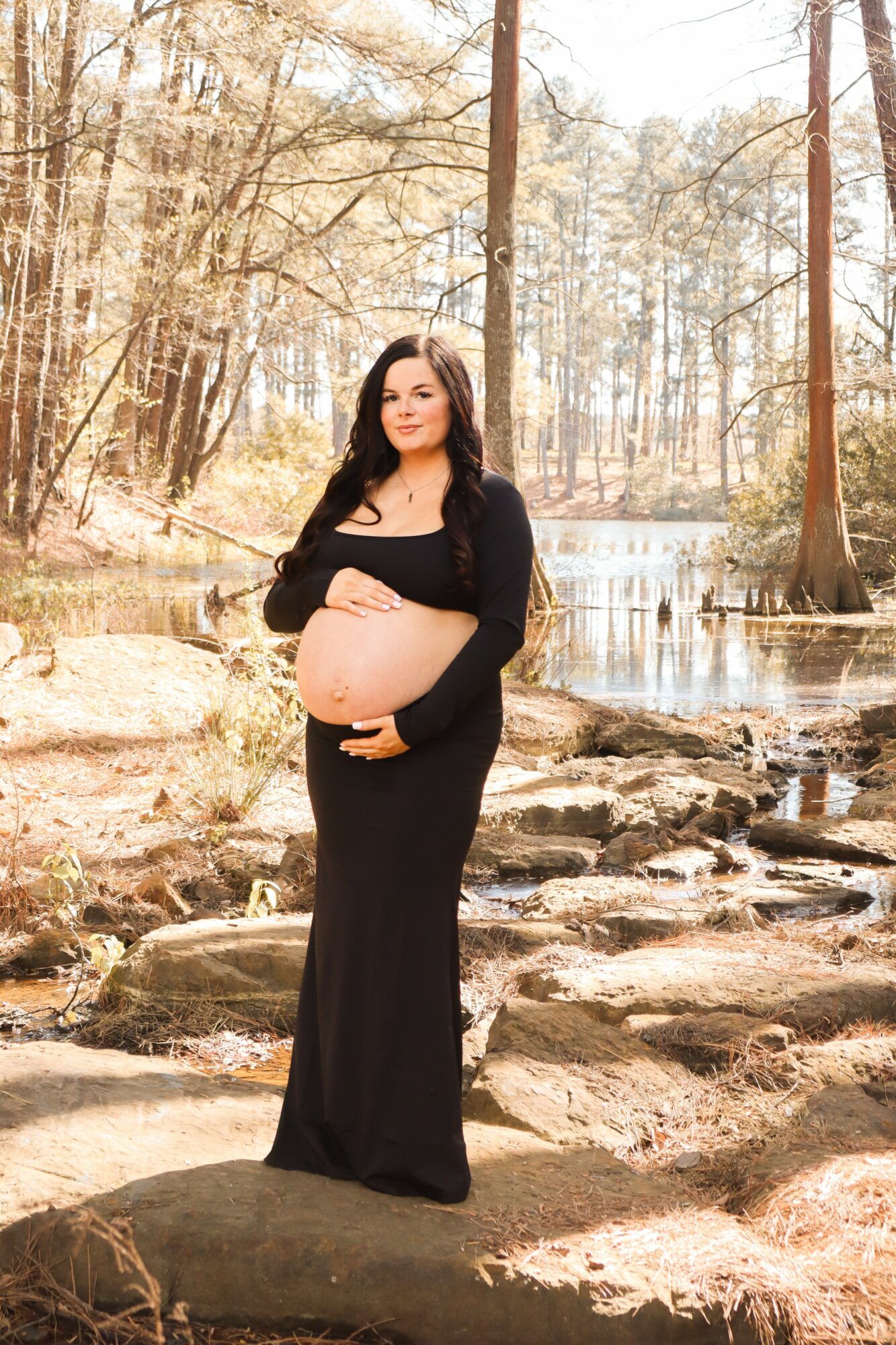 Pregnant woman in a long black dress standing on rocks near a river in a forested area.