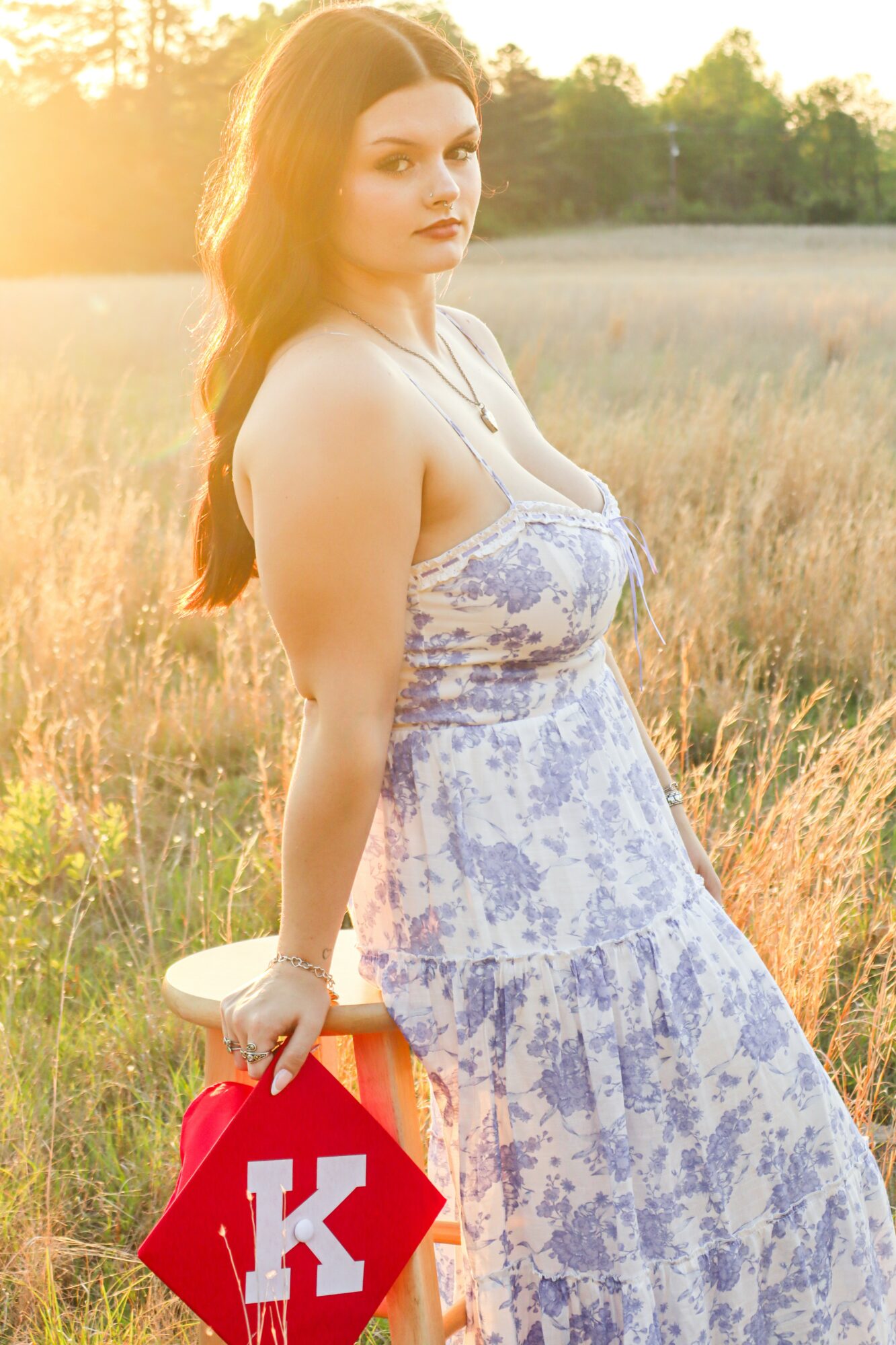 Young woman in a floral dress standing in a field during sunset, holding a red bag with a white letter K.