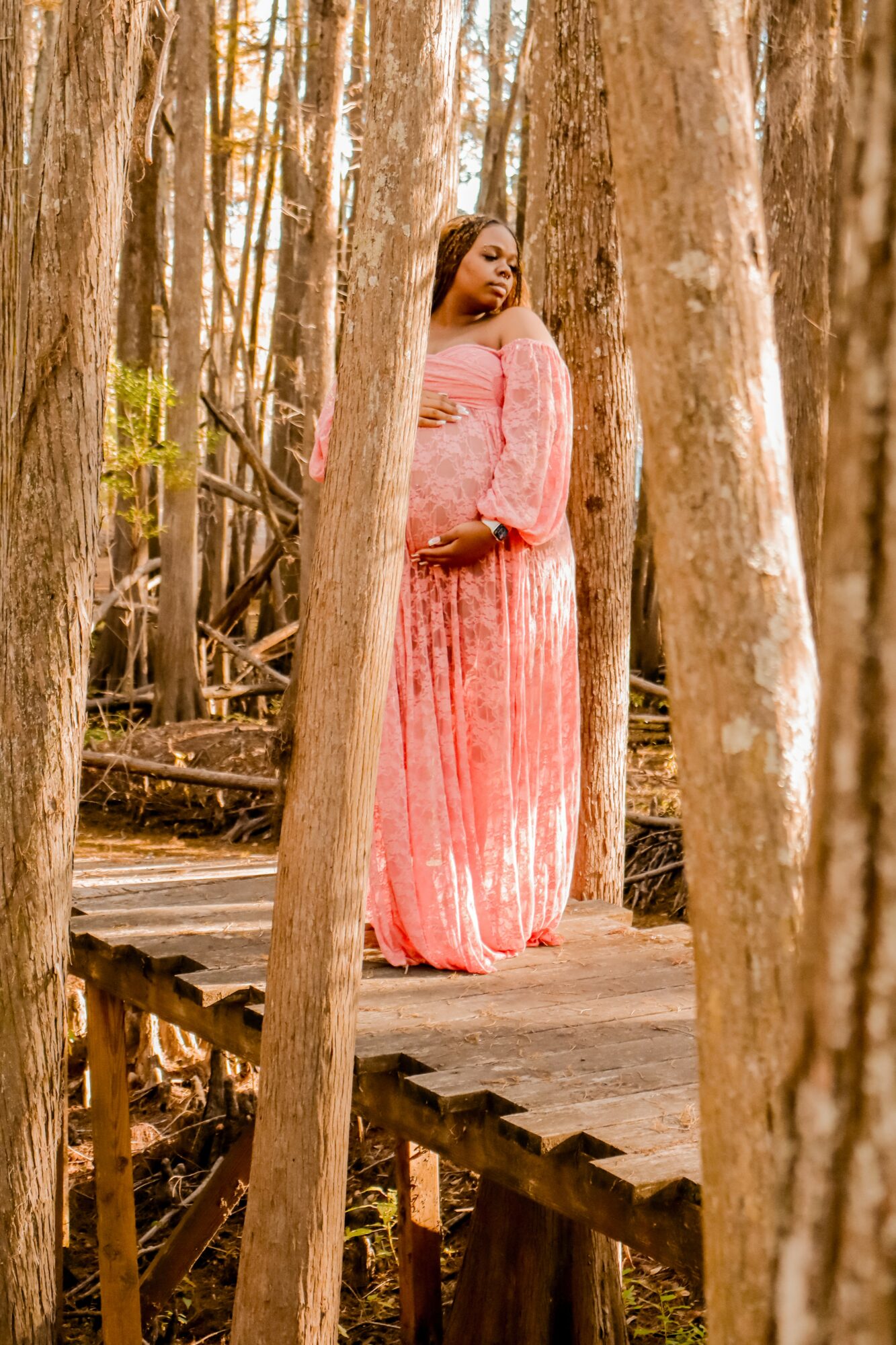 Woman in pink dress standing on wooden bridge in forest, trees surrounding her.
